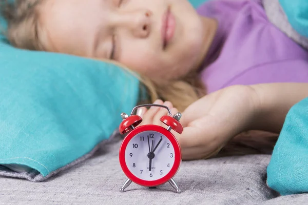 Szép gyermek lány alszik a háttérben, a piros ébresztőóra, amely nyomán neki, reggel. — Stock Fotó