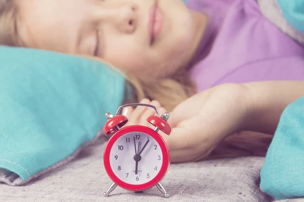 Szép childgirl, a háttérben a piros ébresztőóra, amely nyomán neki, reggel alszik. — Stock Fotó