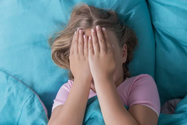Menina loira de cabelos compridos deitada de volta na cama azul cobrindo esconder as mãos do rosto. Acordado cedo. Manhã ensolarada marota. vista superior . — Fotografia de Stock