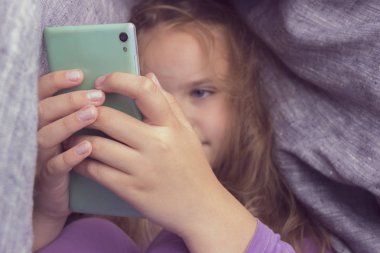 Battaniyenin altında telefon ile oturma sevimli küçük kız.