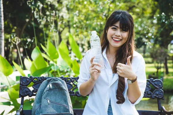 Jovem mulher feliz sentada no banco no parque. Asiático mochileiro menina beber garrafa água relaxamento — Fotografia de Stock