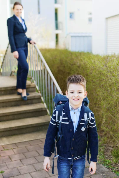 En ung vit pojke går i skolan. Mamma på bakgrund. Stockbild