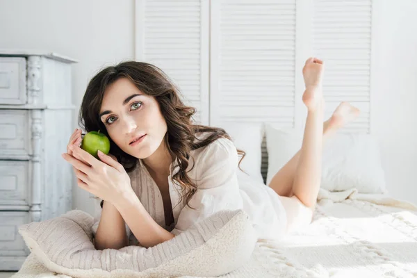 Hermosa chica acostada en la cama y sosteniendo una manzana — Foto de Stock