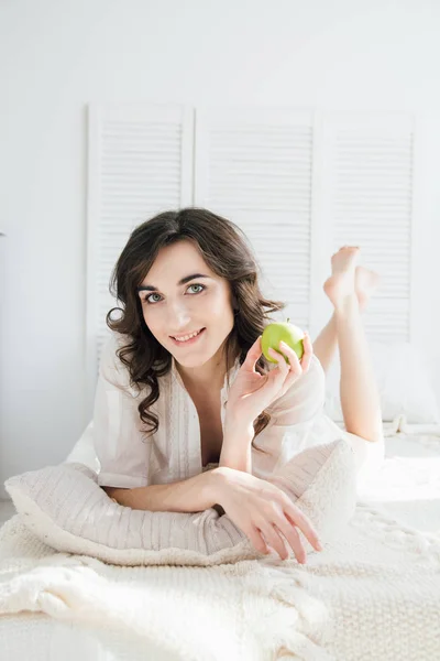 Hermosa chica acostada en la cama y sosteniendo una manzana — Foto de Stock