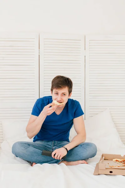 Мужчина смотрит телевизор, сидит на кровати и ест пиццу — стоковое фото