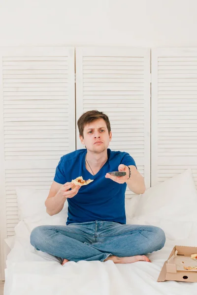 Мужчина смотрит телевизор, сидит на кровати и ест пиццу — стоковое фото