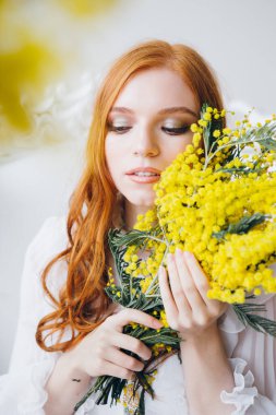 Uzun beyaz elbiseli bir Mimosa 'lı kızıl saçlı güzel bir kızın portresi. Bahar konseri. Tonlama.