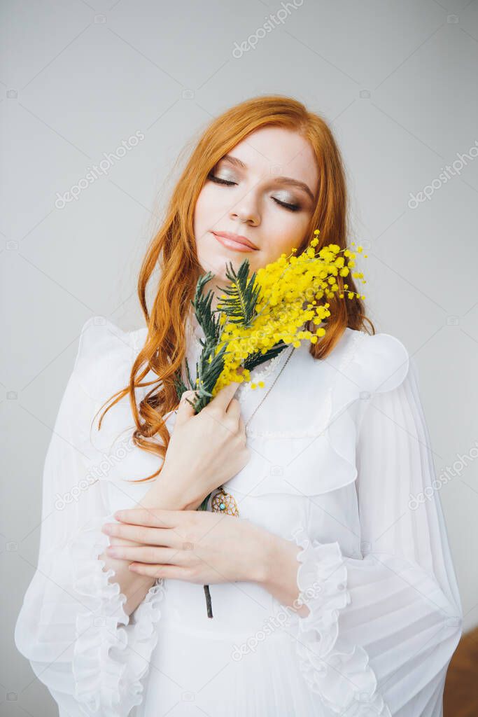 Portrait of a red-haired beautiful girl with a Mimosa in a long white dress. Spring concert. Toning.