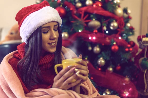 Junge Frau in Weihnachtsmannmütze trinkt Tee oder Kaffee über dem Christbaumhintergrund — Stockfoto