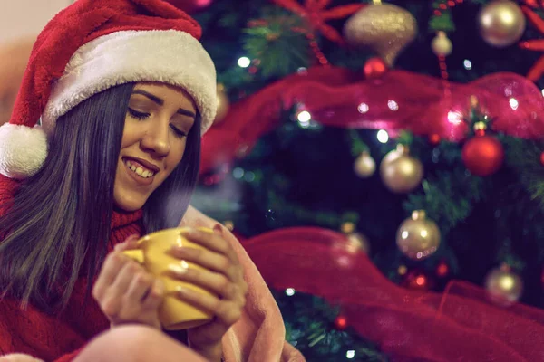 Улыбающаяся девушка в шляпе Санты пьет чай за рождественской ёлкой дома — стоковое фото
