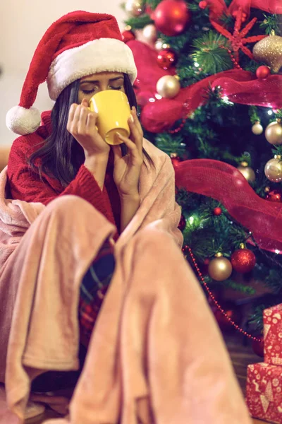 Noel ağacının yanında oturan ve sıcak çay içen bir kadın. — Stok fotoğraf