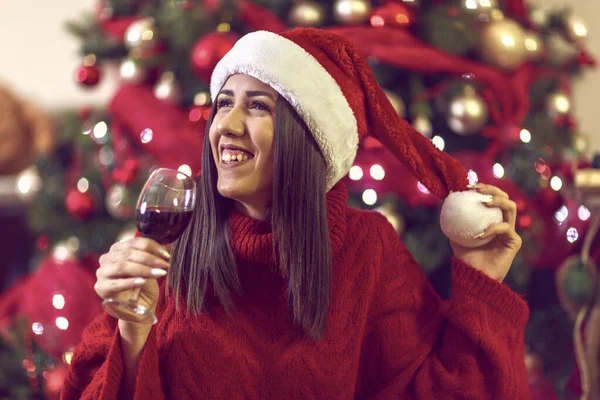 Junge Frau mit Weihnachtsmütze am Weihnachtsbaum zu Hause beim Anstoßen mit Rotwein. — Stockfoto