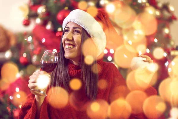 Junge Frau mit Weihnachtsmütze am Weihnachtsbaum zu Hause beim Anstoßen mit Rotwein. — Stockfoto