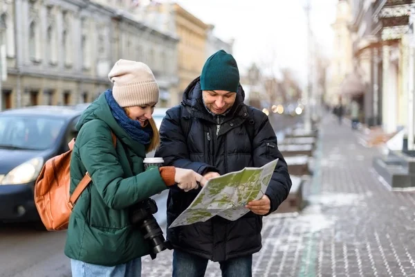 Un par de turistas en el centro histórico de la ciudad están leyendo un mapa mientras visitan la ciudad y se divierten. Chica en ropa de invierno con cámara y taza de café y hombre en ropa de invierno . — Foto de Stock