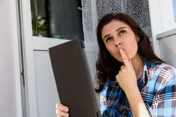 Brunette girl thinking and holding tablet making a selfie or video call in balcony by touching her lips with finger — Stock Photo, Image