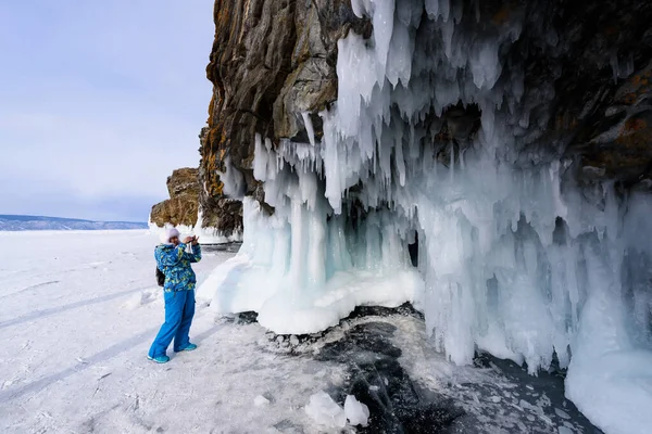 Lago Baikal, Rusia - 10 de marzo de 2020: mujer con chaqueta azul y sombrero blanco toma fotos de rocas heladas con carámbanos en el lago de invierno Baikal — Foto de Stock