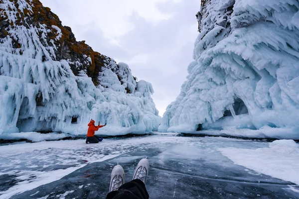 Lake Baikal, Russia - March 9, 2020: Girl in orange jacket and hat takes a selfie against the background of the frozen rocks of lake Baikal in cloudy weather. Feet in ice skates in the foreground — Stock Photo, Image