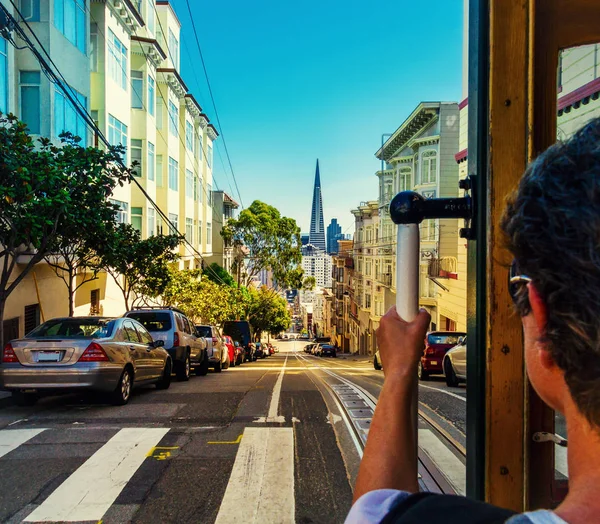 Ride with the cable car in San Francisco. Picture shows a person riding the famous MUNI train on Powell-Mason line down the hill to the bay with the famous landmark of Transamerica Pyramid in front. — Stock Photo, Image