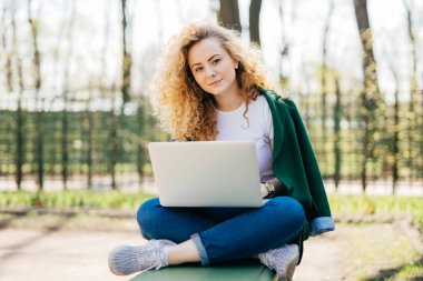 Hafif kıvırcık saçlı ve mavi gözleri açık havada doğrudan kameraya bakarak sevgilisi ile mesajlaşma diz dizüstü tutan bağdaş oturan rahat kıyafetlerle beyaz kadın