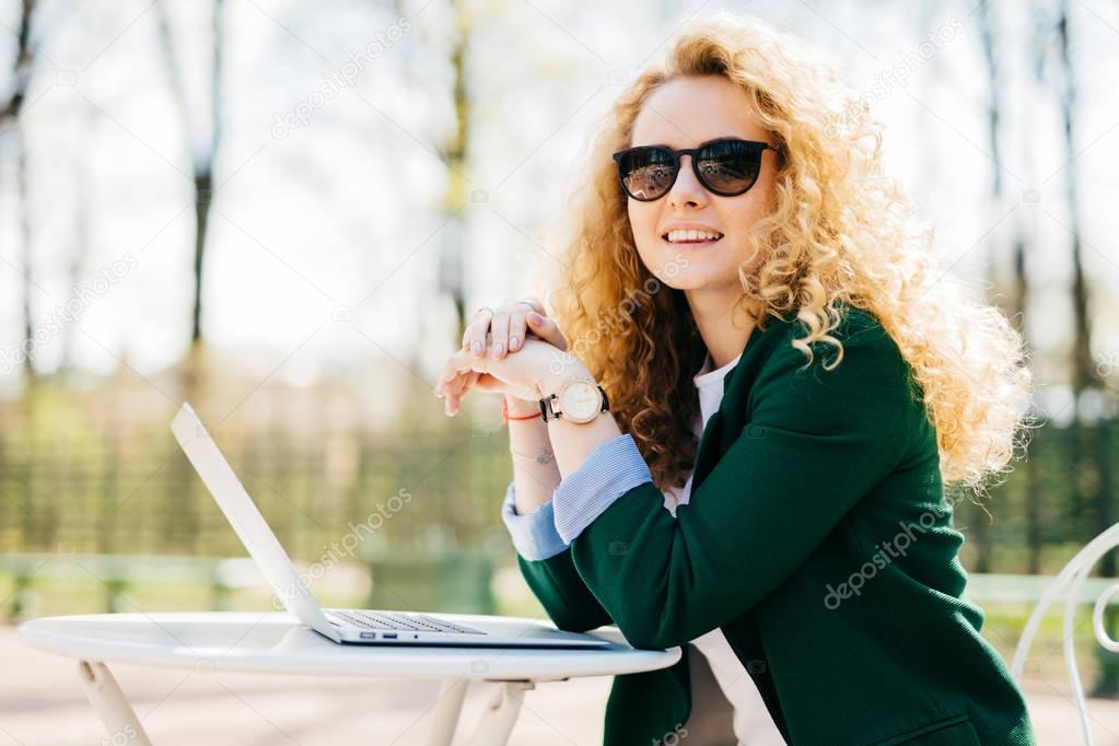 Beautiful young student female with beautiful curly blonde hair elegant dressed using generic laptop computer for writing essay having rest outdoors having charming smile looking into distance