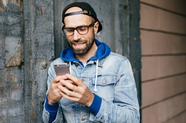 Ücretsiz Internet kullanımı, e-posta kontrol, komik video izlemek, uygulamaları smartphone tutan büyük gözlük takıyor denim anorak sakallı şık erkekte yakışıklı yatay portresi