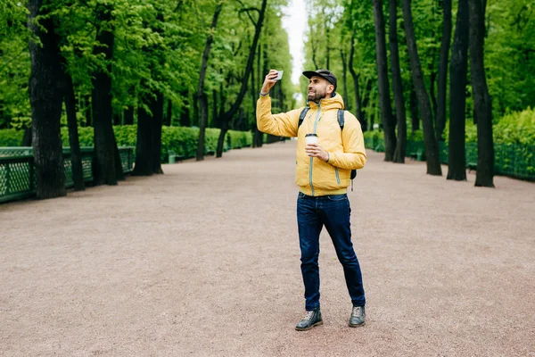 Outdoor portrait of young handsome tourist dressed in stylish clothes holding backpack and takeaway coffee posing into camera of his smartphone making selfie isolated over green nature background — Stock Photo, Image