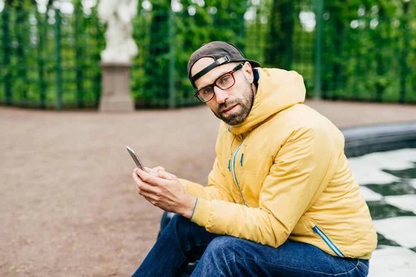 Attractive man with thick beard wearing trendy cap, yellow anorak and glasses sitting outdoors with telephone. Tourist male in casual clothes looking pensively into camera while posing in green park