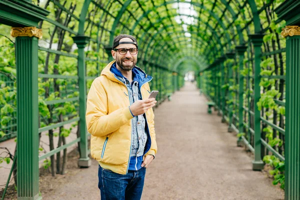 Young student male with beard having stylish clothes standing against green background using mobile phone for downloading video. Bearded hipster in yellow anorak resting outdoors typing messages — Stock Photo, Image
