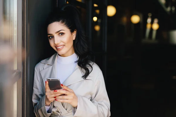 Sorrindo mulher morena bonita com rabo de pônei ter manicure vermelho vestido formalmente segurando smartphone fazendo compras on-line enquanto espera por trem. Empresária com celular indo para o trabalho — Fotografia de Stock