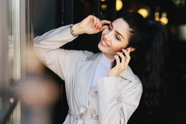 Mulher elegante morena magro com rabo de cavalo vestido com casaco branco com sobrancelhas escuras, olhos, pele pura, manicure agradável conversando sobre telefone celular tocando sua cabeça com a mão olhando sorrindo de lado — Fotografia de Stock