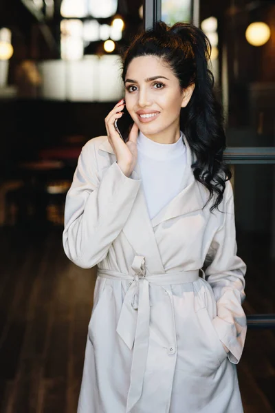 Vertical portrait of elegant brunette businesswoman in white coat communicating over cell phone while standing outdoors. Caucasian woman with beautiful appearance using modern gadget. Fashion concept — Stock Photo, Image