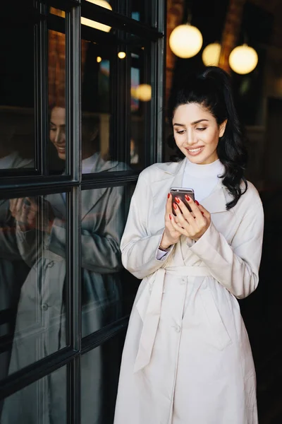 Mulher morena bonita elegante com cauda de pônei vestida com capa de chuva branca com rosto agradável e manicure vermelho perfeito segurando smartphone olhando para a tela com sorriso agradável — Fotografia de Stock