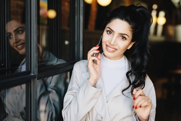 Cute female with wavy dark hair, charming eyes, well-shaped lips and white even teeth wearing white clothes having red manicure communicating over telephone touching her hair with her gentle hand — Stock Photo, Image