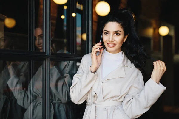 Mujer morena encantadora con el pelo ondulado atado en cola de caballo que tiene buena cara pálida buscando elegantemente mientras posando al aire libre utilizando gadget moderno. Linda joven mujer vestida con ropa de moda — Foto de Stock