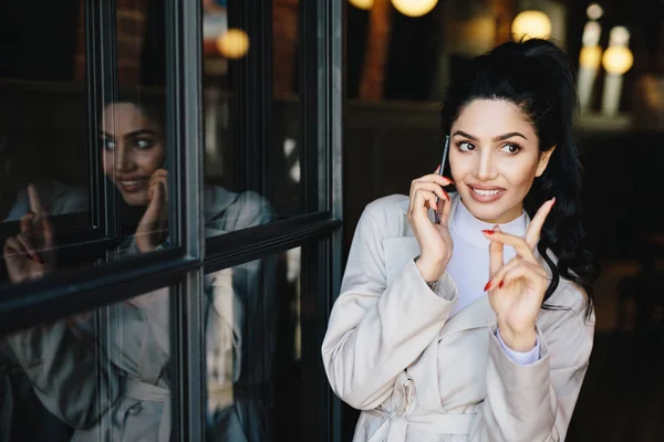 Encantadora joven morena con una sonrisa agradable con manos suaves y hermosa manicura comunicándose por teléfono móvil con su amiga levantando el dedo mientras se hace una buena idea para la fiesta — Foto de Stock