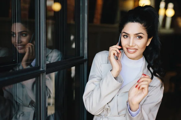 Mooie vrouw met zwarte haren hebben dunne donkere wenkbrauwen, heldere ogen met lange wimpers en volledige goed gevormde lippen dragen van elegante kleding bedrijf smartphone met iemand spreken en glimlachen — Stockfoto