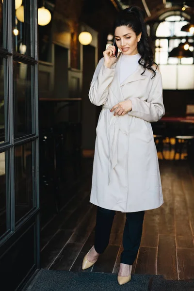 Retrato completo de mulher de negócios elegante vestindo sobretudo branco, sapatos elegantes e calças pretas em pé no café chamando seu parceiro de negócios enquanto espera por ele. Pessoas, conceito de tecnologia — Fotografia de Stock