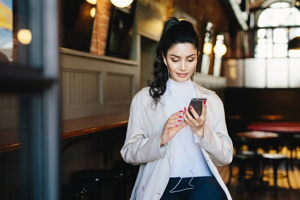 Красивая брюнетка с хвостиком и привлекательным внешним видом, сидя в кафе, используя смартфон серфинг в Интернете, используя бесплатный Wi-Fi соединение и слушая свою любимую музыку с наушниками — стоковое фото