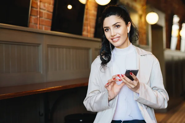 Mulher morena de boa aparência com sobrancelhas finas e olhos quentes vestindo roupas elegantes sentadas no café esperando seu pedido ouvindo áudio livro on-line usando telefone celular com sonhos — Fotografia de Stock