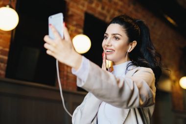 Beyaz ceketli uzun kirpiklerini, dolgun dudaklar ve sağlıklı cilt kulaklık ve yapım selfie ile farklı parçaları dinlerken kafede dinlenme sırasında iyi bir ruh hali olan karanlık gözlere sahip esmer kadın