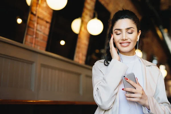 Mulher morena repousante com aparência encantadora fechando os olhos com alegria enquanto desfruta de música alta em seus fones de ouvido. Mulher elegante ouvir sua música favorita, tendo tempo livre e relaxamento — Fotografia de Stock