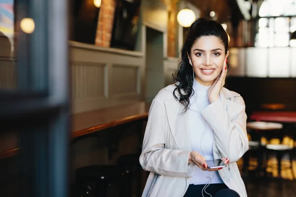 Happy brunette woman with make-up and manicure wearing white jacket holding smartphone and having earphones in ears listening to music or audiobook on earphones. Dreaming female in cafe using mobile