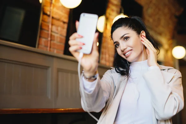 Привлекательная женщина с темными волосами, здоровой кожей и яркими глазами, держащая смартфон в руке, делая селфи и слушая музыку или аудиокнигу во время отдыха в кафе. Люди и концепция отдыха — стоковое фото