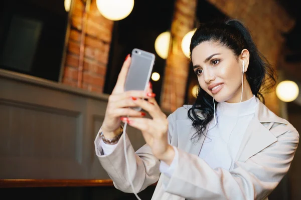 Mulher bonita com cabelo escuro amarrado na cauda do pônei fazendo selfie enquanto sentado no café usando seu smartphone. Mulher bem tratada em roupas elegantes olhando na tela de seu telefone fazendo foto — Fotografia de Stock