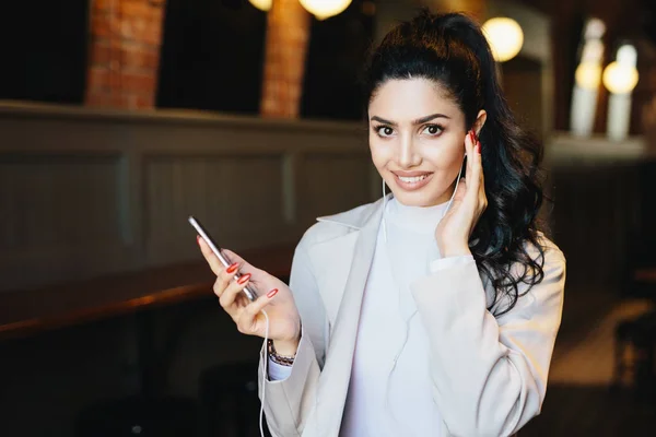 Mujer linda con cola de caballo oscuro con pestañas largas y ojos expresivos sentados en la cafetería escuchando música en línea disfrutando de conexión a Internet de alta velocidad. Concepto de personas, belleza y tecnología — Foto de Stock