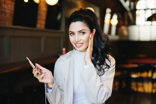 Morena modelo joven con aspecto encantador que tiene manos delicadas vestidas con chaqueta blanca escuchando sus pistas favoritas en los auriculares, utilizando la aplicación de música en línea en el teléfono celular en el acogedor restaurante — Foto de Stock