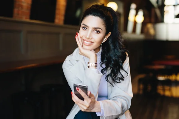 Mulher branca jovem morena com cauda de pônei longa com rosto bonito e mãos gentis com manicure vermelha ouvindo sua música favorita com fones de ouvido e telefone celular posando no fundo do café — Fotografia de Stock