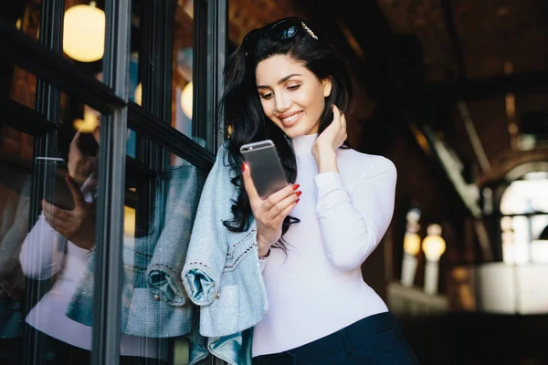 Jovem empresária morena em roupas elegantes em pé contra o interior do café esperando que as pessoas tenham reunião usando telefone inteligente dentro de casa, mensagens de texto on-line. Conceito de comunicação — Fotografia de Stock