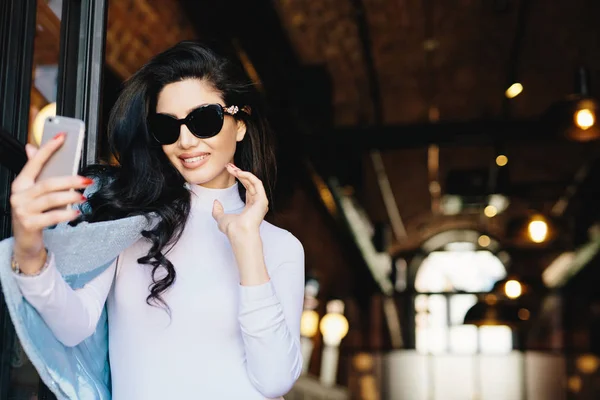 Hymyilevä glamouria nainen aurinkolasit, valkoinen pusero ja takki poseeraa kameran hänen älypuhelin samalla selfie valokuvaus itse. Onnellinen tyylikäs malli poseeraa kahvilassa matkapuhelimella — kuvapankkivalokuva