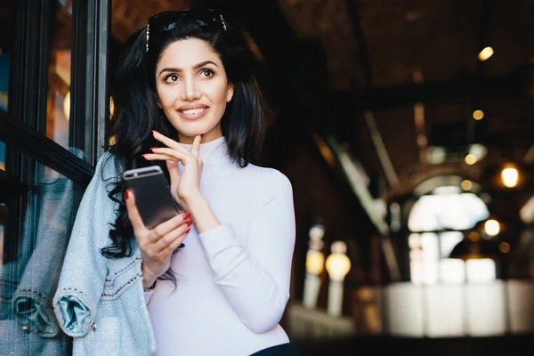 Portret van doordachte glamour vrouw met donker haar dragen zonnebril en witte formele kleding surfen sociale netwerken met behulp van smartphone genieten van online communicatie poseren tegen café interieur — Stockfoto